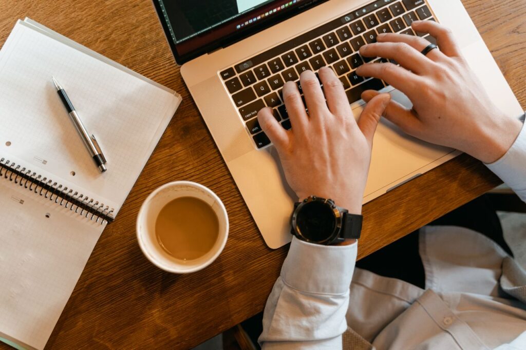 Hands typing on a laptop next to a cup of coffee and a notebook on a wooden table.
