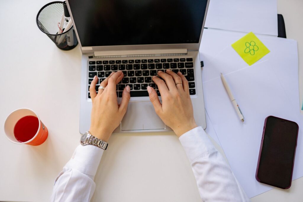 A person working on a laptop in a bright workspace with a drink and smartphone nearby.